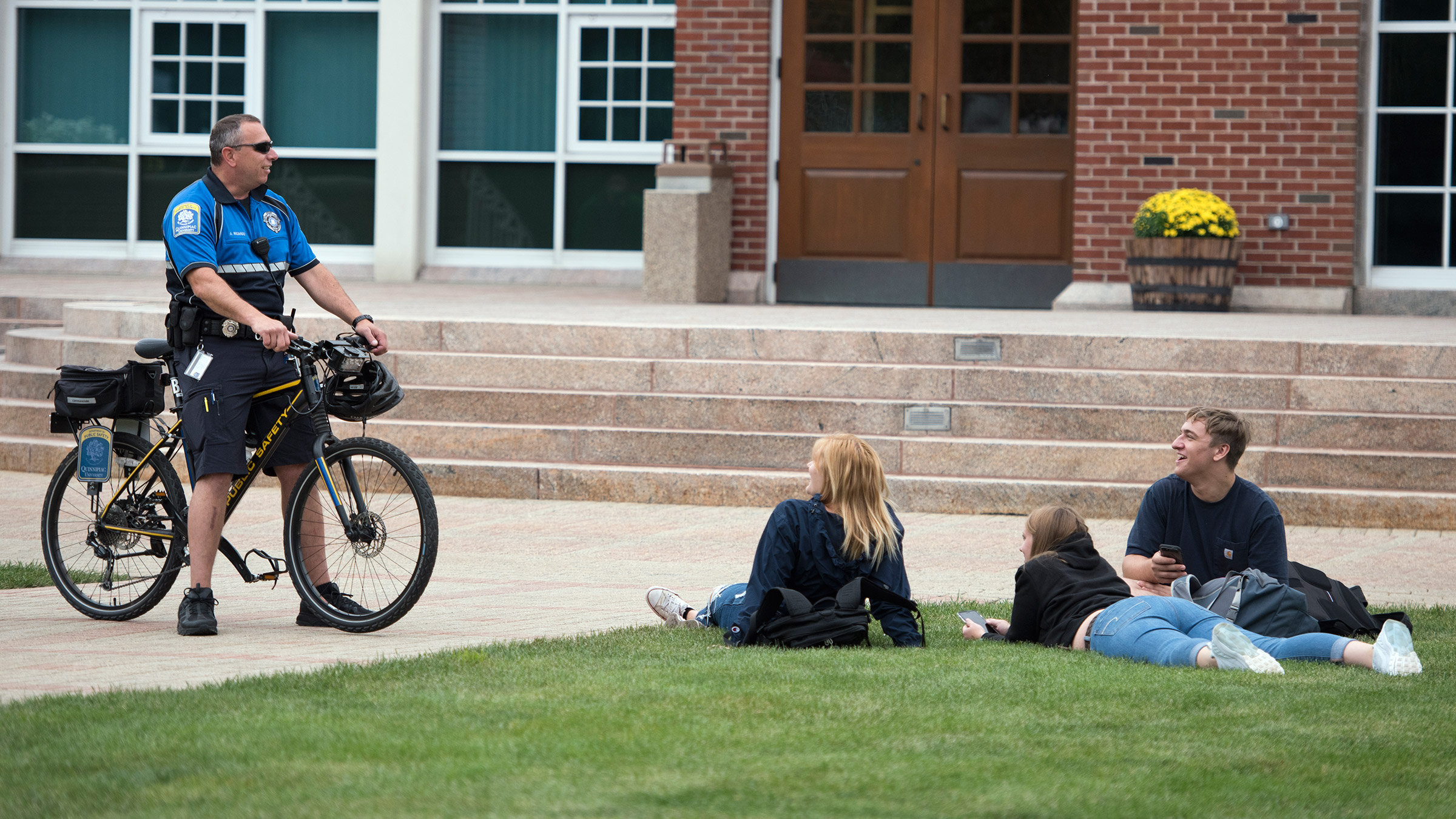 A public safety officer stands with his patrol bike and talks to students lying in the grass on the quad.